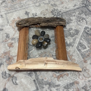 Pode incluir: Uma moldura retangular de madeira com um painel central de vidro transparente. A moldura é decorada com um pedaço de madeira flutuante escura e desgastada no topo e uma peça mais clara na base. Um arranjo em forma de flor de pedras lisas cinzentas e castanhas está fixado no centro do vidro.
