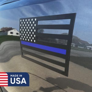 May include: Black metal American flag emblem with a blue line, featuring white stars and stripes. The emblem is affixed to a vehicle's surface. The words "MADE IN USA" are printed on a blue and red sticker.