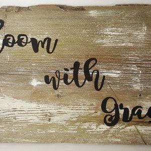 May include: A rustic wooden sign with the words "Bloom with Grace" written in black cursive lettering. The sign is decorated with two burlap flowers and twine.