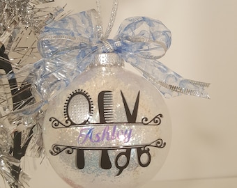 May include: A white glitter Christmas ornament with a blue ribbon and a silver bow. The ornament has a black silhouette of a comb, scissors, and a mirror with the name "Ashley" written on a banner.