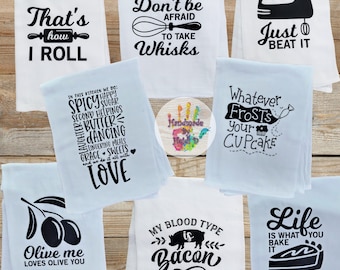 May include: Ten white tea towels with black text and graphics. The towels feature various kitchen-themed sayings, including "That's how I roll", "Don't be afraid to take whisks", "Just beat it", "In this kitchen we do: spicy, happy, sugar, second helpings, butter, dancing, lingering meals, grace, sweets, and we do it all with love", "Olive me loves olive you", "My blood type is bacon", "Whatever frosts your cupcake", and "Life is what you bake it".