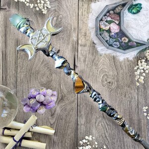 May include: A silver and gold wand with a triple moon design at the top. The wand is decorated with crystals and a tiger's eye stone. The wand is lying on a wooden surface with a small pile of amethyst stones and a crystal ball.
