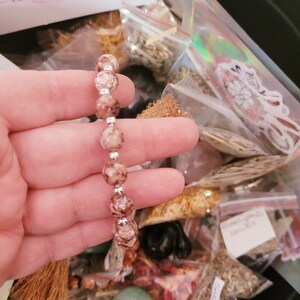 May include: A close-up of a pink and white beaded bracelet with silver accents. The bracelet is being held by a hand, and the background is a variety of small bags filled with different items.