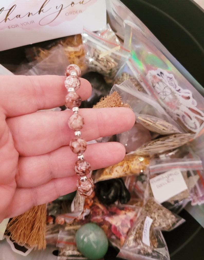 May include: A close-up of a pink and white beaded bracelet with silver accents. The bracelet is being held by a hand, and the background is a variety of small bags filled with different items.