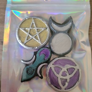 Pode incluir: Cinco pequenos amuletos redondos de resina com vários símbolos pagãos. Os amuletos são pintados em ouro, prata, azul-esverdeado, roxo e branco. Os símbolos incluem um pentagrama, uma lua crescente, um triskel e uma lua crescente estilizada.