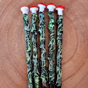 Peut inclure: Cinq stylos noirs avec des motifs en spirale vert et jaune et des embouts en forme de champignon rouge et blanc. Les stylos sont disposés en rangée sur une surface en bois. Le texte "Witch Aroma UK" est visible sur la surface.