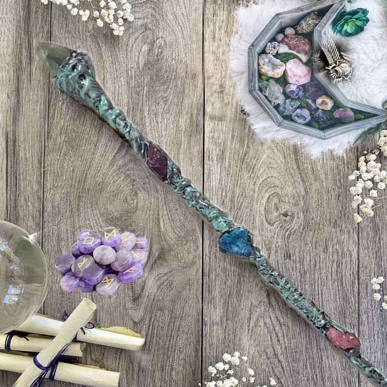 May include: A green, blue, and purple crystal wand with a wooden handle. The wand is decorated with crystals and runes. The wand is lying on a wooden surface with a small pile of purple crystals and a rolled up scroll.