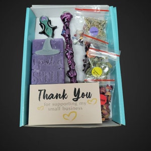 Könnte beinhalten: Eine türkisfarbene Geschenkbox mit einer lila und weißen Seifenstange, auf der "I cast a spell" steht. Die Box enthält außerdem einen lila und goldenen Zauberstab, eine Dankeskarte und drei kleine Beutel mit getrockneten Kräutern, die mit "Lavender", "Lemon Balm" und "Rose" beschriftet sind.