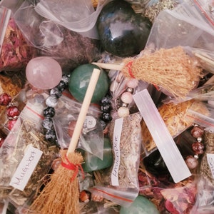 May include: A collection of various herbs, crystals, and other magical supplies in small clear plastic bags. The items include dried herbs, crystals, and a small wooden broom. The bags are labeled with the names of the herbs, such as "Mugwort" and "Lavender".