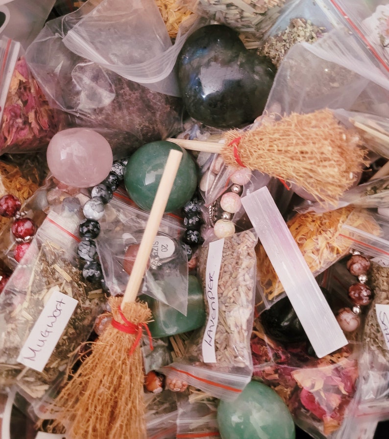 May include: A collection of various herbs, crystals, and other magical supplies in small clear plastic bags. The items include dried herbs, crystals, and a small wooden broom. The bags are labeled with the names of the herbs, such as "Mugwort" and "Lavender".