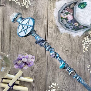 May include: A blue and silver wand with a white pentagram at the top. The wand is decorated with various crystals and stones. The wand is lying on a wooden surface with other crystals and stones scattered around it.