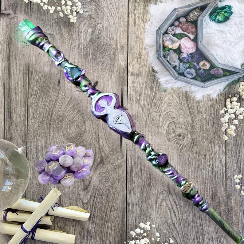 May include: A purple and green wand with a silver and white goddess symbol, surrounded by amethyst crystals and scrolls.