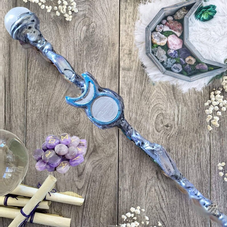 May include: A blue and silver wand with a moon and star design. The wand has a clear crystal ball on top and is surrounded by purple crystals, scrolls, and other decorative items.