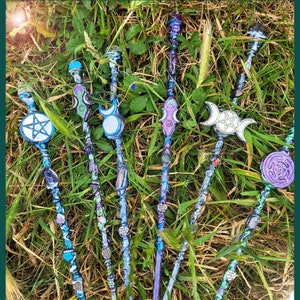 May include: A collection of six decorative wands with various designs, including a pentagram, a crescent moon, and a triple moon. The wands are made of wood and decorated with colourful paint, glitter, and crystals.
