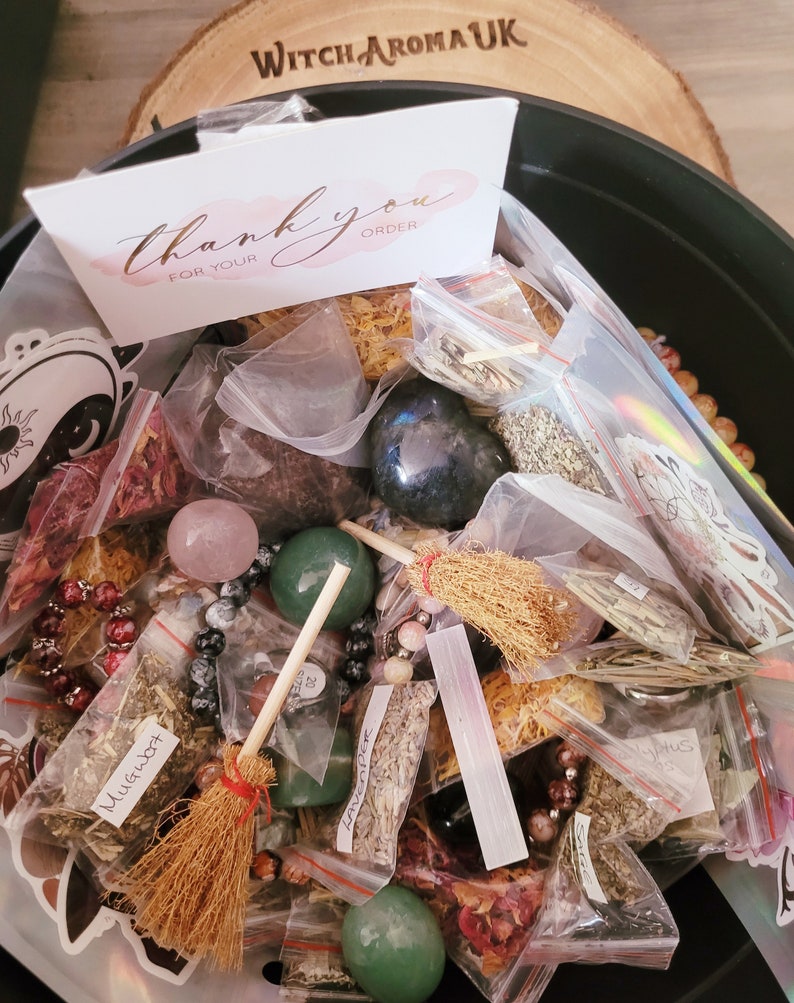 May include: A black cauldron filled with various herbs, crystals, and other magical ingredients. The cauldron is surrounded by a wooden surface with the text "Witch Aroma UK" engraved on it. A small card with the text "Thank you for your order" is visible in the top left corner of the image.