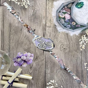 May include: A wooden wand with a purple and silver design, featuring a circular symbol with three spirals. The wand is surrounded by amethyst crystals, scrolls, and a wooden box filled with crystals.