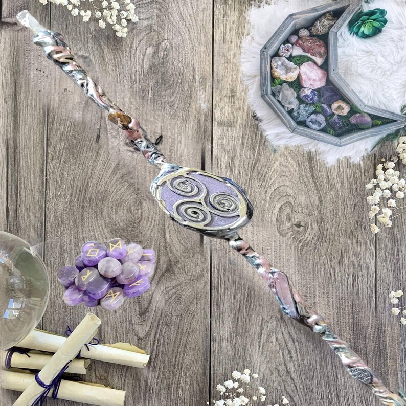 May include: A wooden wand with a purple and silver design, featuring a circular symbol with three spirals. The wand is surrounded by amethyst crystals, scrolls, and a wooden box filled with crystals.