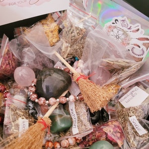 May include: A black container filled with various items, including small bags of herbs, crystals, and beads. The bags are labeled with names like "Sage", "Lavender", and "Eucalyptus Leaves". There are also two small brooms and a thank you note for an order.