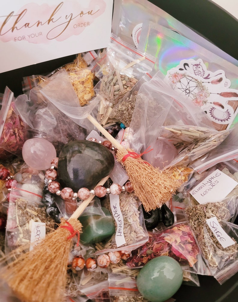 May include: A black container filled with various items, including small bags of herbs, crystals, and beads. The bags are labeled with names like "Sage", "Lavender", and "Eucalyptus Leaves". There are also two small brooms and a thank you note for an order.