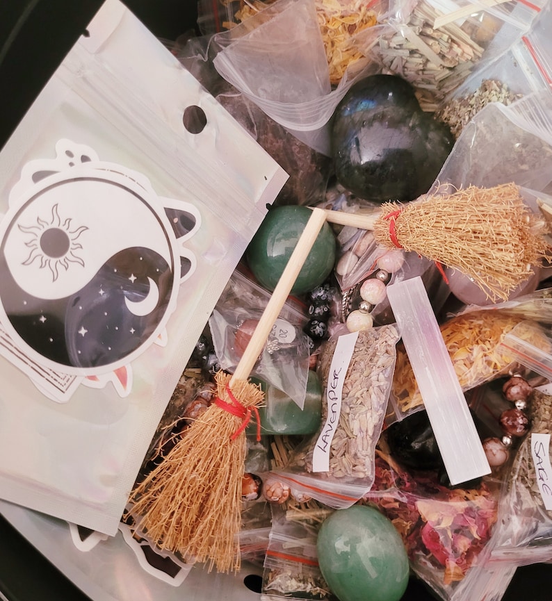 May include: A collection of various herbs, crystals, and other magical ingredients in a black container. The items are arranged in small plastic bags and include a small broom, a yin yang symbol sticker, and a green crystal. The bags are labeled with the names of the herbs, such as "Lavender" and "Sage".