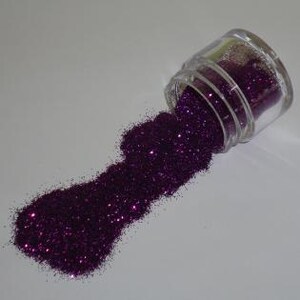 May include: A small, clear glass jar tipped over, spilling a stream of sparkling, dark purple glitter. The glitter forms a textured pile on a white surface, catching the light and creating a shimmering effect. The jar's lid is visible.