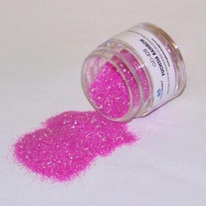 May include: A clear, small jar of fuchsia glitter is tipped over, spilling a pile of the sparkly material onto a white surface. The jar's label reads "Fuchsia Rainbow" and "GD-428". The glitter appears to be fine and reflective.