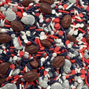 May include: A close-up of a mix of decorative sprinkles in red, white, silver, and navy blue. The sprinkles include football shapes, round white sprinkles, silver skull shapes, and red and navy blue rod-shaped sprinkles.