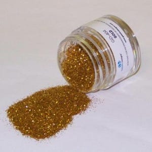 May include: A clear glass jar labeled "GD-404 GOLD" is tipped over, spilling gold glitter onto a white surface. The glitter is a fine, sparkling powder, creating a shimmering effect. The jar is partially filled, and the label includes additional text and a logo.