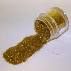 May include: A small, clear glass jar tipped over, spilling a stream of gold glitter onto a white surface. The glitter sparkles, catching the light. The jar is partially filled with the same shimmering gold material.