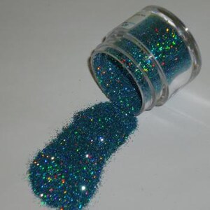 May include: A small, clear jar tipped over, spilling teal glitter onto a white surface. The glitter has a holographic effect, reflecting multiple colors. The jar is partially in focus, with the glitter forming a small pile.