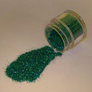 May include: A small, clear plastic jar tipped on its side, spilling out a pile of vibrant green glitter. The glitter sparkles with hints of blue and reflects light. The jar is partially filled with the same glitter, suggesting a craft supply or decorative element.