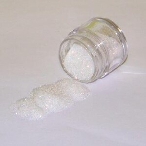 May include: A small, clear glass jar tipped on its side, spilling a pile of iridescent white glitter. The glitter sparkles, reflecting light. The jar has a clear plastic lid. The background is a plain white surface.