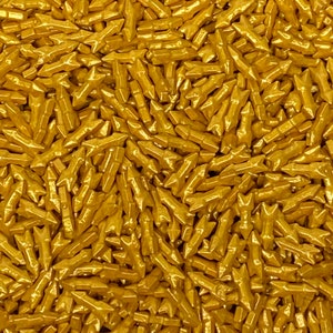 May include: Close-up of a pile of gold-colored, arrow-shaped sprinkles. The sprinkles are small and have a metallic sheen, creating a textured, glittering effect. These decorative sprinkles are ideal for adding a festive touch to baked goods.