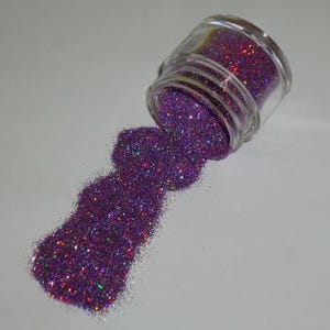 May include: A small, clear glass jar tipped over, spilling a pile of vibrant purple glitter. The glitter has a sparkling, iridescent quality, reflecting multiple colors. The jar is partially filled, and the glitter forms a textured mound on the surface.