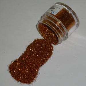 May include: A small, clear glass jar tipped over, spilling out a pile of bronze glitter. The glitter sparkles, catching the light. The jar has a label with text. The background is a plain, light surface.