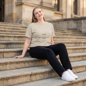 Puede incluir: Una camiseta beige de manga corta con el texto "FACT" dentro de un recuadro rectangular sobre las palabras "YOU'RE ABSOLUTELY RIGHT!" en letras negras, combinada con vaqueros negros y zapatillas blancas.