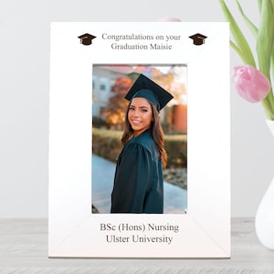 May include: A white photo frame displaying a graduation photograph. The frame is inscribed with "Congratulations on your Graduation Maisie" and "BSc (Hons) Nursing Ulster University". A vase of pink tulips stands beside it.