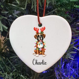 May include: Heart-shaped white ceramic Christmas ornament with a Boxer dog wearing reindeer antlers and Christmas lights. The dog's name, "Charlie," is written in black script below the dog. A red string is attached for hanging.
