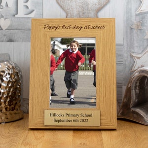 May include: A light oak picture frame with the inscription "Poppy's first day at school". The frame contains a photo of a child in a red top and grey shorts. A gold plaque at the bottom reads "Hillocks Primary School September 6th 2022".
