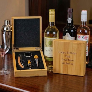 May include: A bamboo wine accessory set in a wooden box with a black foam insert containing a corkscrew, a bottle stopper, and a drip ring. The box lid is engraved with the text Happy Birthday Dad Love from Amber xx, next to three wine bottles.