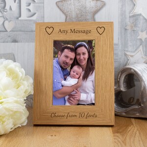 May include: A wooden photo frame with a light brown finish. The frame has a rectangular opening for a photo and a small heart on each side of the top edge. The text "Any Message" is engraved on the top edge of the frame. The text "Choose from 10 fonts" is engraved on the bottom edge of the frame. A family photo is displayed in the frame.
