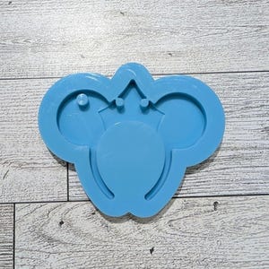 May include: A light blue silicone mold in the shape of a cartoon character's head with large ears and a crown. The mold has a smooth surface and is designed for crafting. The mold is on a light wood grain surface.