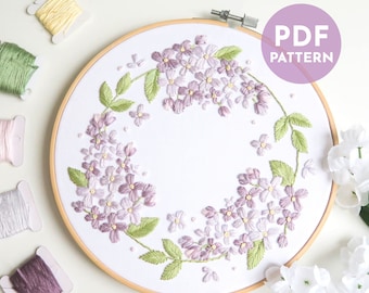 May include: A lilac flower embroidery pattern in a hoop with a white background. The pattern includes a wreath of lilac flowers and leaves. The text "PDF PATTERN" is in a purple box in the upper right corner of the image.