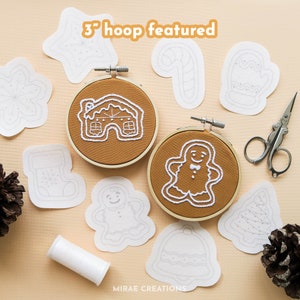 May include: A collection of 3-inch embroidery hoops featuring various Christmas designs, including a gingerbread house, a gingerbread person, a stocking, a candy cane, a mitten, a bell, a Christmas tree, and a star. The hoops are on a light brown surface with white embroidery floss and pine cones.