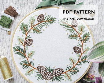 May include: A PDF pattern for an embroidery hoop wreath featuring pine branches and pine cones. The wreath is in shades of green and brown with gold accents. The text "PDF PATTERN INSTANT DOWNLOAD" is at the top of the hoop.