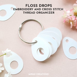 May include: White oval-shaped floss bobbin holders with a hole in the center. The bobbins are attached to a silver ring. The text "FLOSS DROPS EMBROIDERY AND CROSS STITCH THREAD ORGANIZER" is above the bobbins. The bobbins are on a pink surface.