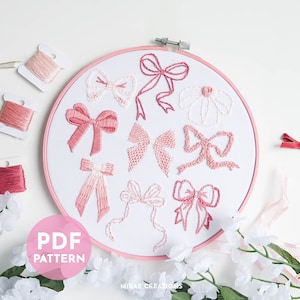 Pode incluir: Um aro de bordado rosa com um fundo de tecido branco com vários laços bordados em rosa e branco. O aro é rodeado por flores brancas e uma fita rosa.
