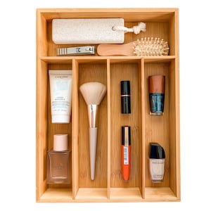 May include: A bamboo drawer organizer with six compartments. The compartments contain a variety of beauty products, including a foot file, nail clippers, a makeup brush, a bottle of perfume, a tube of hand cream, a lipstick, a nail polish, and a bottle of nail polish remover.
