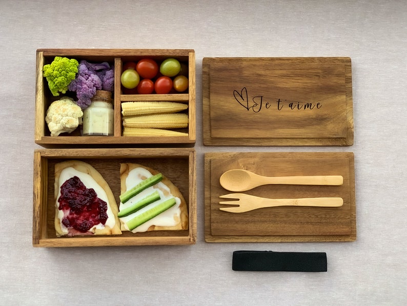 K&ouml;nnte beinhalten: Eine Lunchbox aus Holz mit zwei F&auml;chern. Das obere Fach enth&auml;lt Blumenkohl, Brokkoli, Mais, Tomaten und ein kleines Glas Dip. Das untere Fach enth&auml;lt zwei St&uuml;cke Fladenbrot mit Belag. Die Lunchbox wird mit einem Tablett aus Holz mit L&ouml;ffel und Gabel, einer Holzkiste mit der Gravur "Je t'aime" und einem schwarzen Band geliefert.