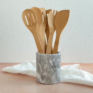 May include: A set of wooden kitchen utensils in a gray marble utensil holder. The utensils include a slotted spoon, a solid spoon, a spatula, and a fork.